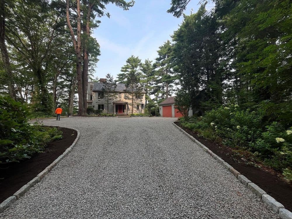 Gravel driveway leading to a large house surrounded by trees and greenery.