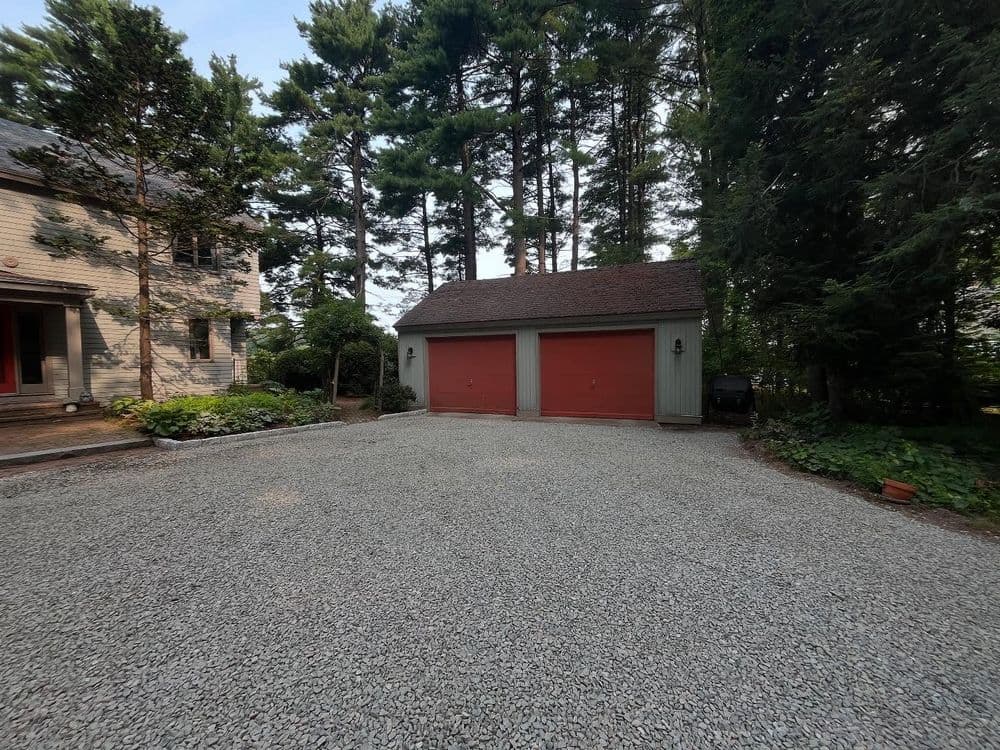 Gravel driveway leading to a two-car garage surrounded by trees.